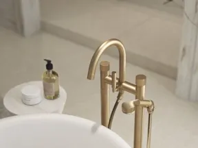 Modern gold freestanding bathtub faucet and white tub with bath products on a side table in a minimalist bathroom.