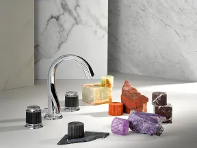 Chrome faucet surrounded by colorful mineral and stone samples on a marble surface with marble backdrops