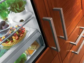 Open refrigerator door revealing fresh herbs, fruits, and a fruit tart inside wooden kitchen cabinets.