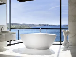 Modern bathroom with freestanding white bathtub and large windows overlooking ocean and hills.