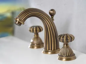 Antique brass bathroom faucet with ornate design and dual handles mounted on a white sink against gray wall.