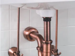 Copper plumbing pipes and fittings under a white sink with hot and cold water valves against tiled wall.