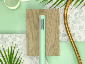 Mint green handheld shower head with gold hose on wood and terrazzo surfaces with green leaves