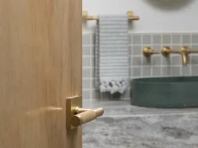 Partially open wood door with brass handle showing modern bathroom with round mirror and green sink.