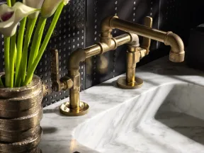 Brass industrial faucet on white marble sink with black soap and gold vase holding calla lilies