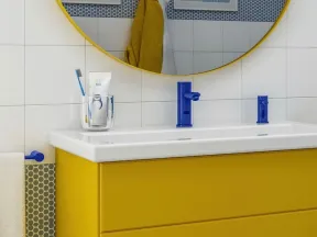 Modern bathroom sink with yellow vanity, blue faucet, round mirror, and hexagonal tile backsplash.