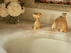 Elegant bathroom sink with gold faucet, ornate mirror, and a vase of white flowers on marble countertop.