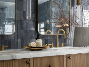 Modern bathroom vanity with brass fixtures, marble countertop, dark tiled backsplash, and decorative vase with branches.