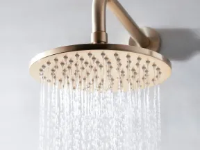 Brushed metal rainfall showerhead with water flowing against a light gray wall background.