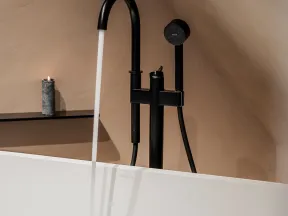 Modern black freestanding bathtub faucet running water with a lit candle on a beige wall shelf.