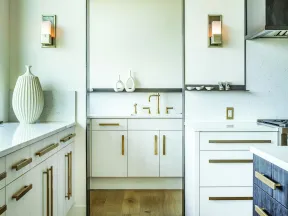 Modern kitchen with white cabinetry, gold handles, wooden floor, and decorative vases by the sink area.