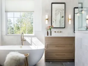Bright modern bathroom with freestanding tub, wooden vanity, rattan stool with fur, and patterned rug.
