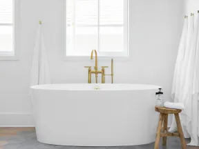 Modern white freestanding bathtub with gold faucet under a window, white towels, and wooden stool on hex tile floor.