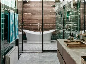 Modern bathroom with green tiles, hexagonal floor, freestanding tub, glass shower, and wooden vanity with gold fixtures.