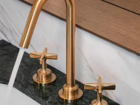 Modern gold faucet with running water on dark green marble countertop and wooden backsplash.