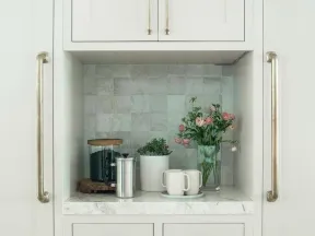 Minimalist kitchen nook with marble countertop, white cabinets, coffee accessories, plants, and a vase of flowers.