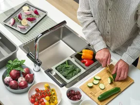 Person slicing zucchini on wooden board with various fresh vegetables and kitchen sink nearby in modern kitchen
