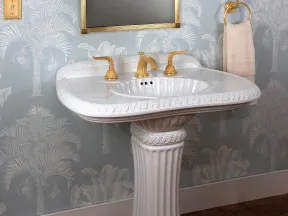 Elegant white pedestal sink with gold fixtures against blue palm tree wallpaper and wooden floor.