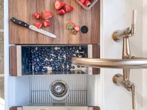 Wooden cutting board with sliced strawberries, bins of blueberries, raspberries, and blackberries in a white kitchen sink with brass faucet.