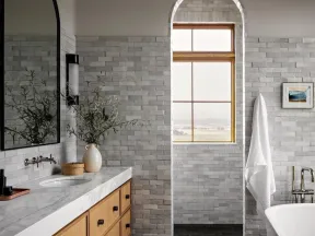 Modern bathroom with marble countertop, wood cabinetry, arched window, freestanding tub, and gray brick walls.