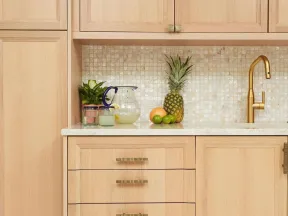 Modern kitchen with light wood cabinets, mosaic tile backsplash, gold faucet, and fresh fruit on white countertop.