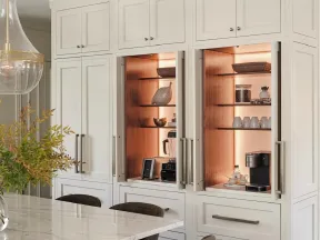 White kitchen cabinets with built-in warm-lit shelves displaying decor and coffee appliances beside a marble countertop.