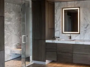Modern bathroom with wooden cabinets, marble countertops, illuminated mirror, and glass shower with marble tiles.