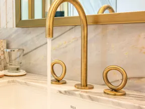 Modern gold bathroom faucet with circular handles on marble countertop and running water in a white sink.