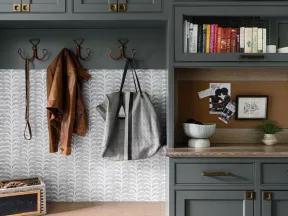 Organized mudroom with gray cabinets, wooden bench, coat hooks, handbags, books, and storage bins below bench.