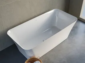 Modern white freestanding bathtub on concrete floor next to wicker basket with rolled towels in bright bathroom.