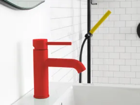 Modern bathroom sink with a bold red faucet against white subway tile walls and a yellow shower handle.