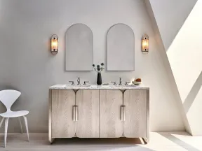 Modern bathroom vanity with dual sinks, arched mirrors, wall sconces, and minimalist white chair in natural light.