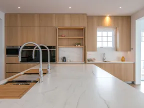 Modern kitchen with expansive marble island, light wood cabinetry, dual sink faucets, and built-in oven