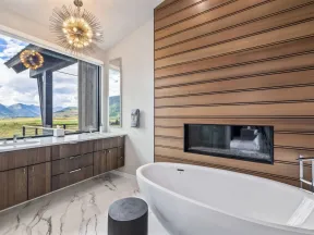 Modern bathroom with freestanding tub, wood panel fireplace, marble floor, large window, and double vanity.