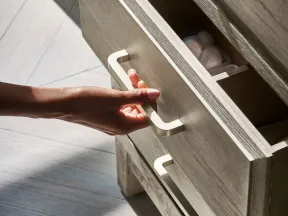 Hand opening a wooden drawer with beige handles showing interior storage and sunlight on wood floor.