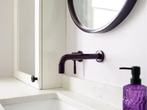 Modern white bathroom sink with wall-mounted dark faucet and purple soap dispenser on marble countertop.