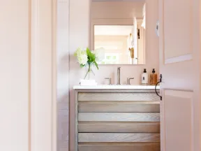 Light wood bathroom vanity with double faucets, mirror, white flowers in vase, and soap dispensers.