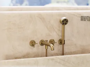 Modern brass bathtub faucet and handheld shower mounted on beige marble surface with soft natural background.