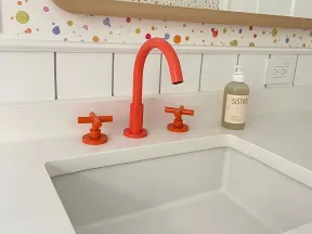 Modern white bathroom sink with vibrant orange faucet and handles, colorful dotted wallpaper, and liquid soap dispenser.