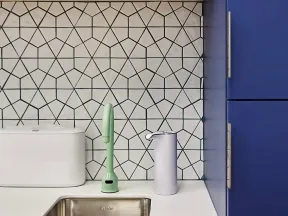 Modern kitchen sink area with geometric tile backsplash, green faucet, white dispenser, and blue cabinets.
