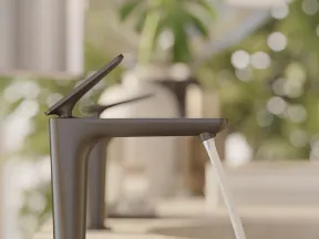 Modern matte black faucet releasing water over a stone sink with blurred green plant background.