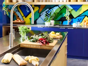 Modern kitchen with stainless steel sink, fresh vegetables, bread, colorful backsplash, and wooden shelves.