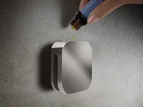 Hand drops essential oil from a brown bottle onto a sleek silver diffuser on a textured gray surface.
