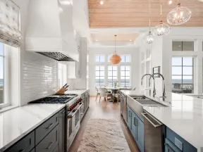 Bright modern kitchen with marble countertops, blue cabinetry, wooden ceiling, and ocean views through large windows.