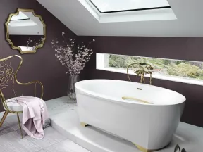 Modern bathroom with white freestanding tub, gold fixtures, skylight, decorative chair, and floral accents.