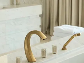 Modern gold bathroom faucet and handheld shower on white marble bathtub with folded towels in background.