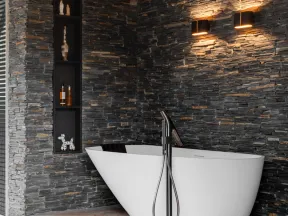 Modern bathroom with freestanding white tub, black stone wall, built-in shelves, and warm wall sconces lighting