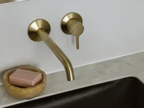 Modern brass wall-mounted faucet over marble countertop with brown sink and stone soap dish with pink soap.
