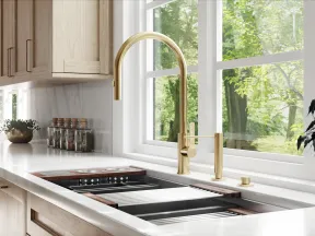 Modern kitchen sink with gold faucet, wooden cabinets, white countertop, and large window overlooking greenery.
