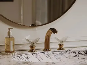 Marble bathroom sink with gold faucet and crystal butterfly handles, round mirror and liquid soap dispenser.